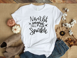 White short sleeve graphic tee with bold black script reading Never Dull Your Sparkle on the front.