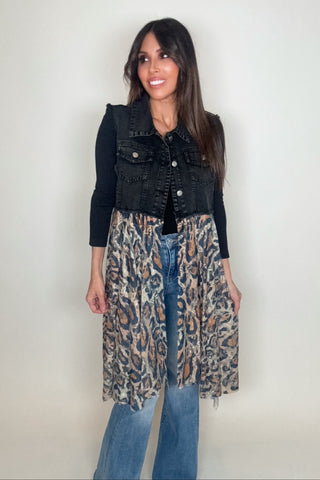 Woman wearing a black denim vest over a leopard print dress with blue jeans on a plain background