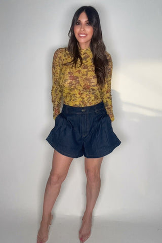 Woman wearing a yellow patterned top and dark blue shorts against a plain background