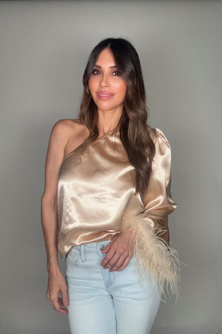 Woman wearing a gold satin top with feather details and light blue jeans against a gray background