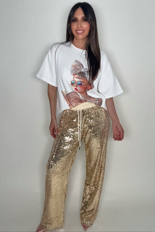 Woman wearing a white t-shirt with a graphic design and gold sequin pants on a plain background