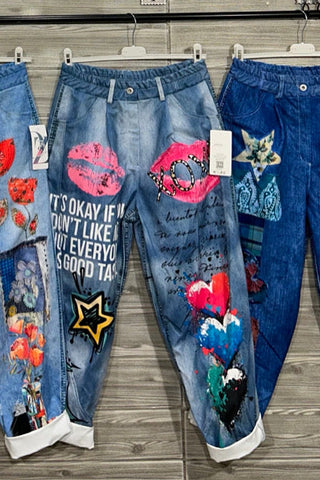 Blue denim joggers decorated with bold pink lips, black XOXO script, star accents, and colorful painted hearts, styled with rolled cuffs.